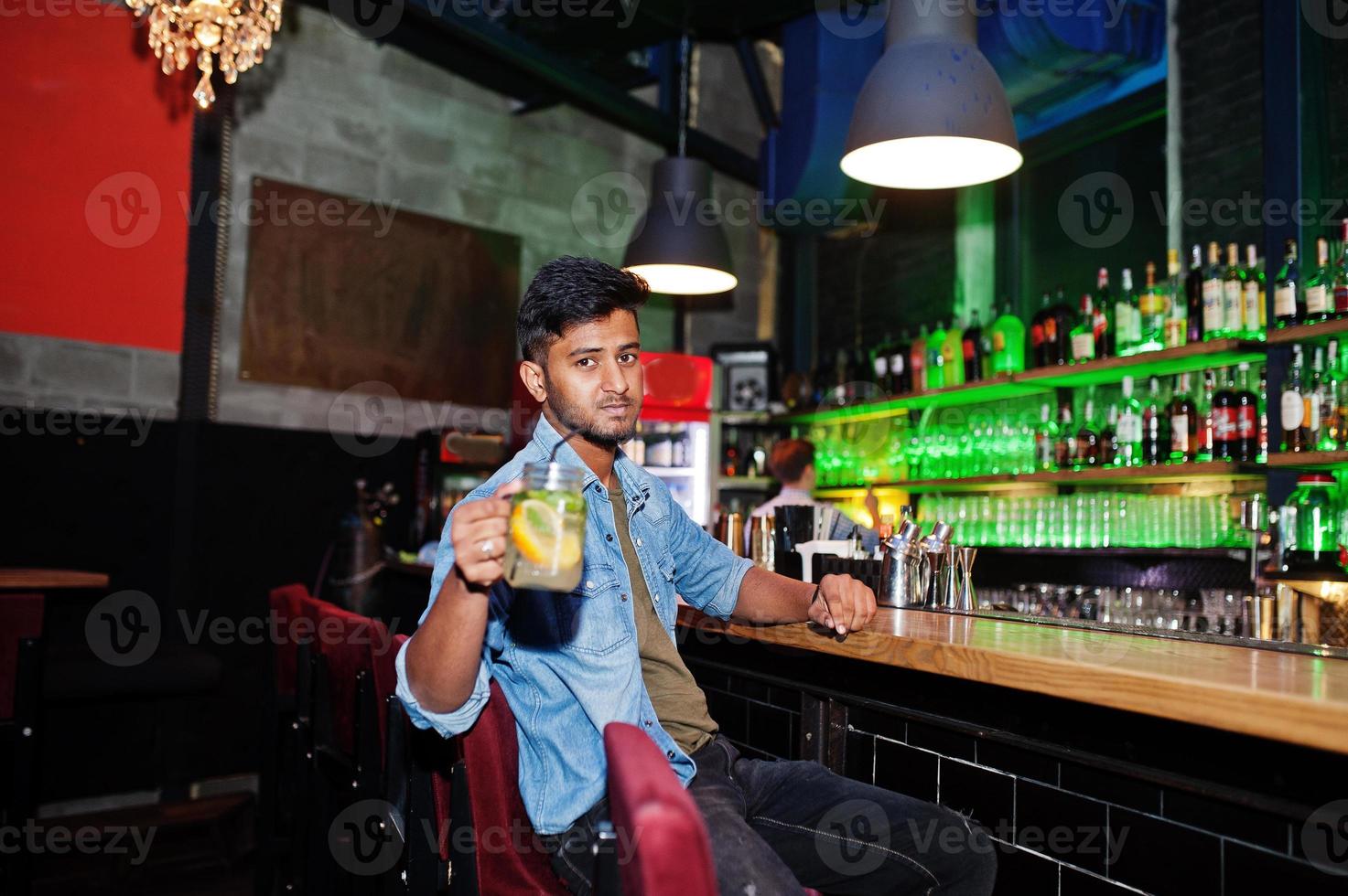 portrait d'un beau sud-asiatique barbu réussi, jeune pigiste indien en chemise de jeans bleu assis dans une boîte de nuit contre un comptoir de bar avec un cocktail et se reposer. photo