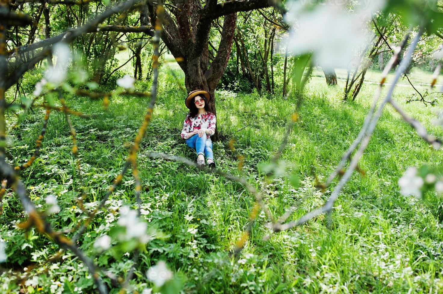 portrait de printemps d'une jeune fille brune à lunettes roses et chapeau au jardin de fleurs vertes. photo