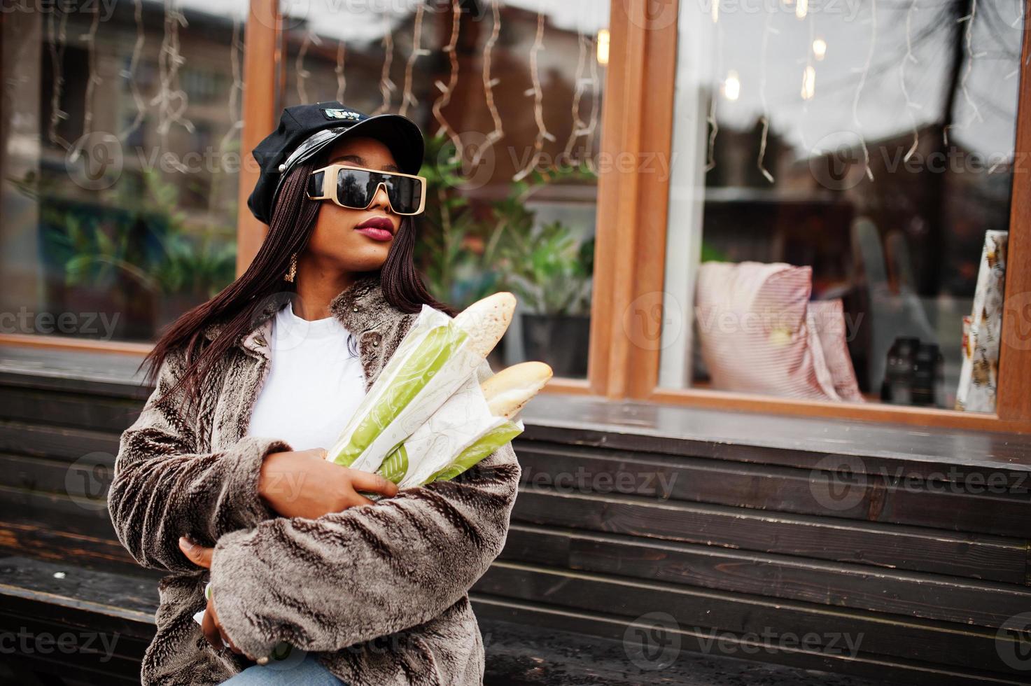 femme afro-américaine à la mode en casquette noire, lunettes de soleil et fourrure avec deux petits pains de baguette à portée de main. photo