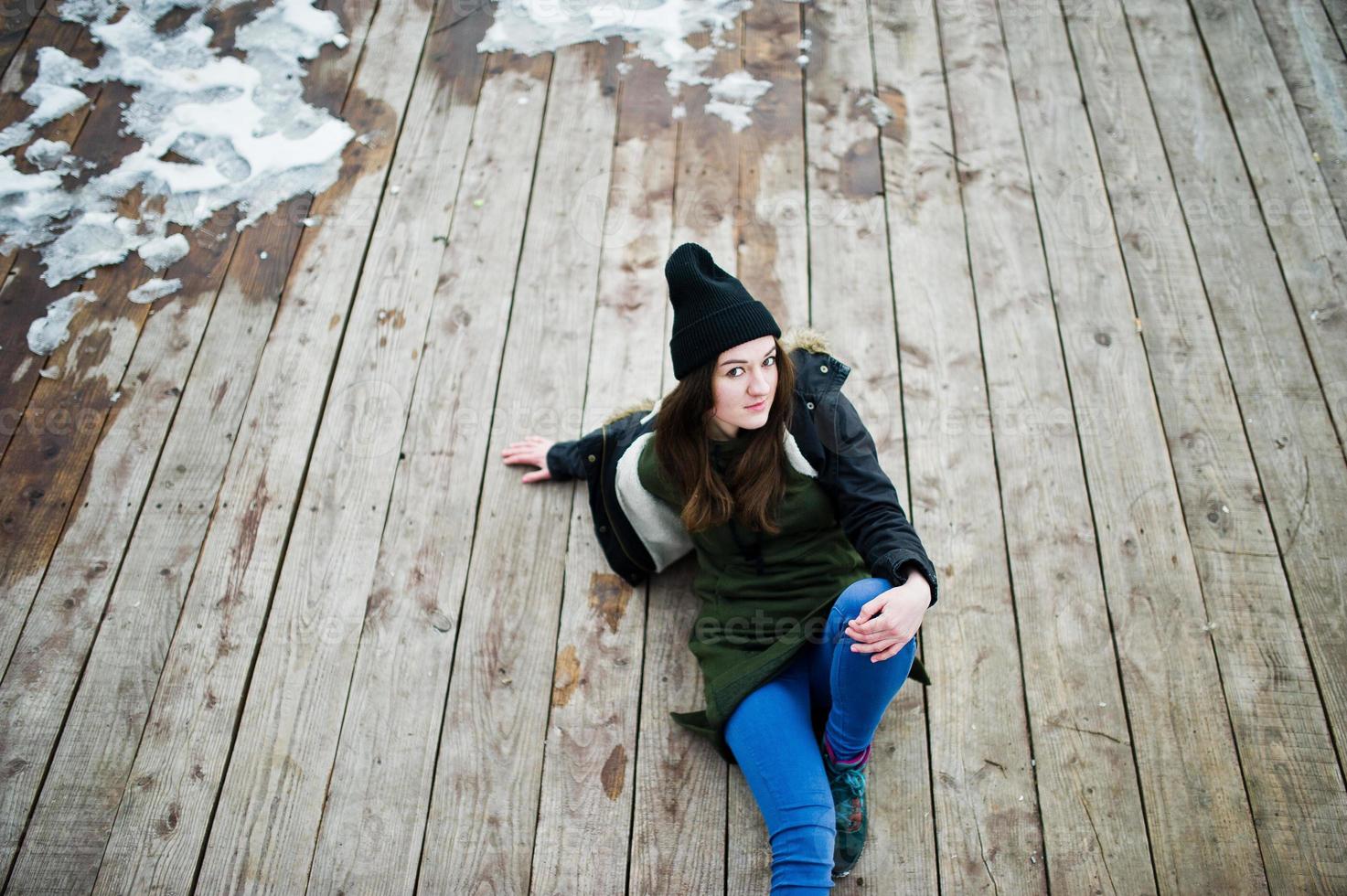 jeune fille porte un long sweat-shirt vert, un jean et un couvre-chef noir assis sur un plancher en bois avec de la neige. photo