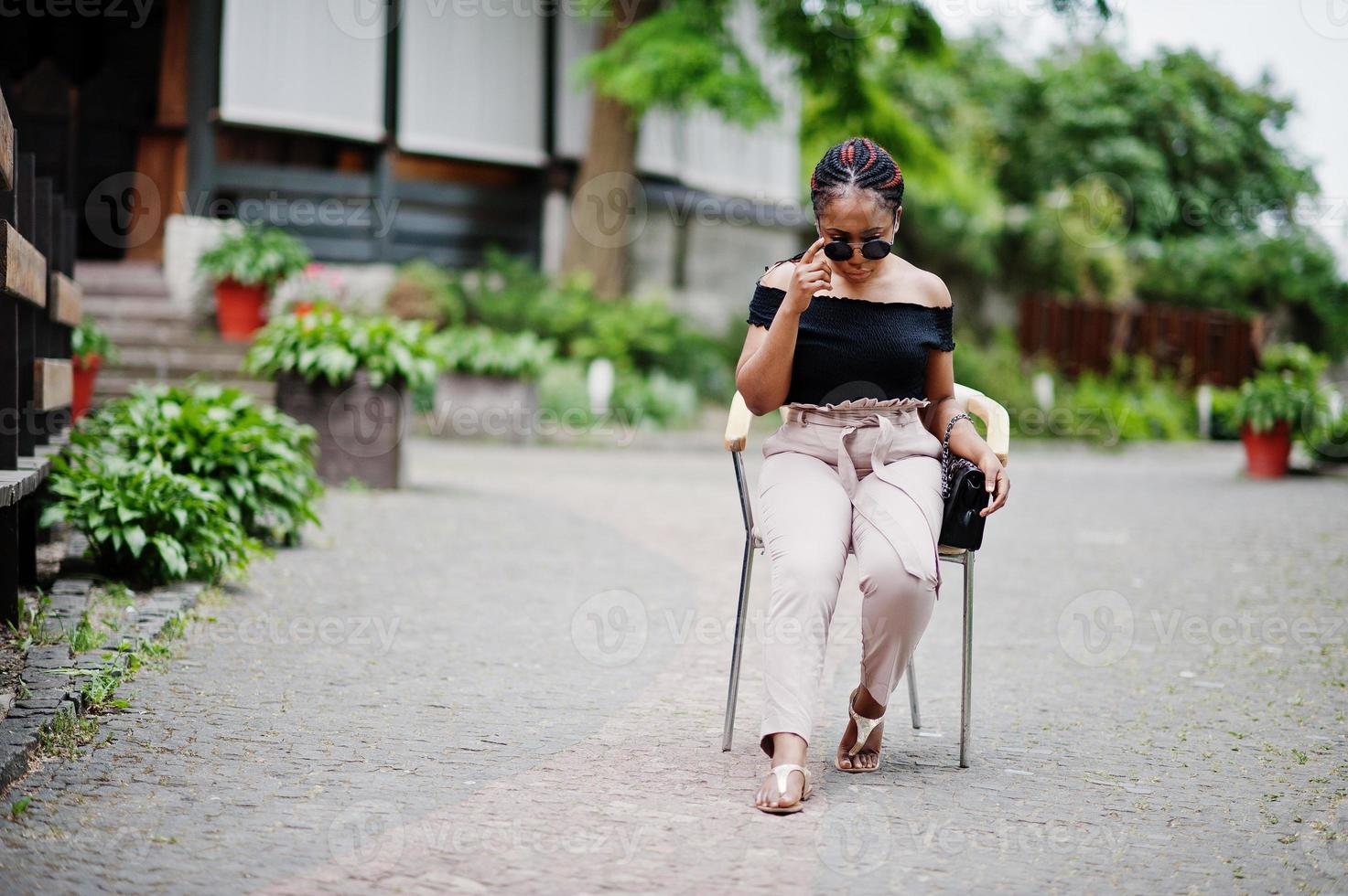 élégante femme afro-américaine à lunettes de soleil posée en plein air sur une chaise. photo