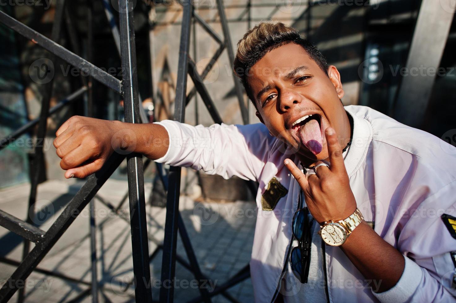 homme arabe hipster élégant posé en plein air dans la rue. le chanteur de rap de style montre la tonque et deux doigts. photo