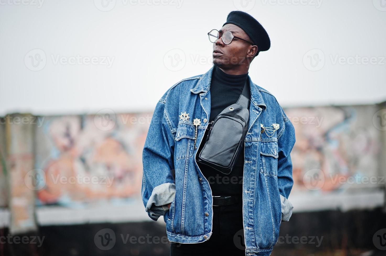 homme afro-américain en veste de jeans, béret et lunettes contre un mur de graffitis sur un toit abandonné. photo