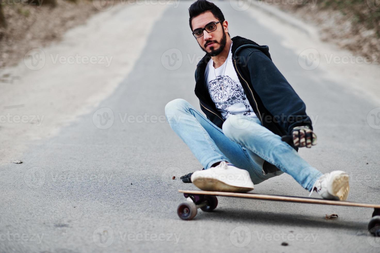 homme arabe de style de rue à lunettes avec longboard longboard sur la route. photo