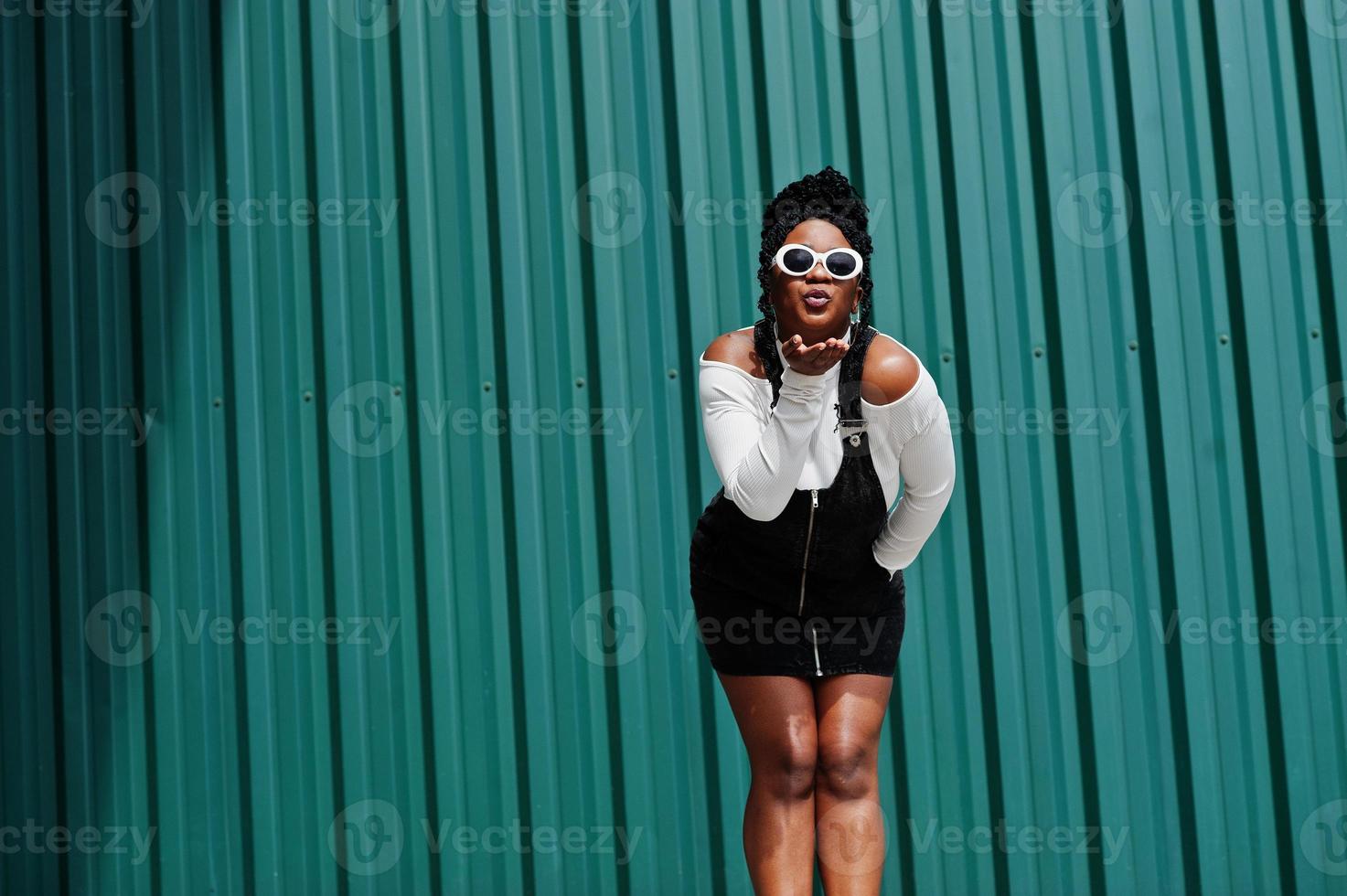 femme africaine en jupe salopette en jean, lunettes de soleil blanches posées contre un mur d'acier vert, envoyant un baiser aérien. photo