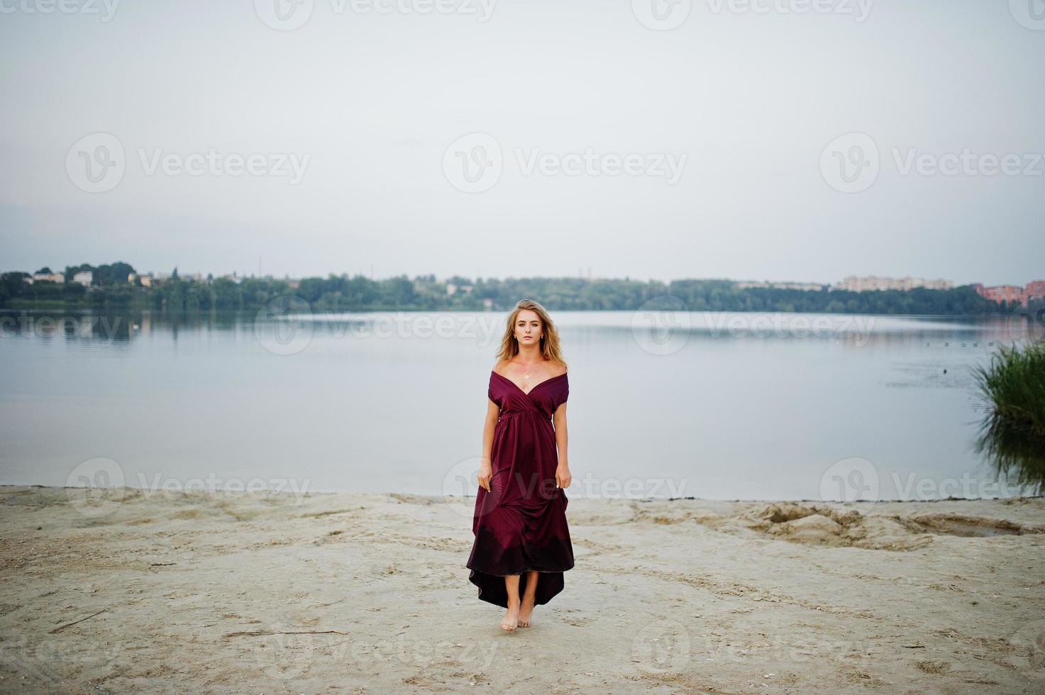 femme aux pieds nus sensuelle blonde en robe de marsala rouge posant contre le lac sur le sable. photo