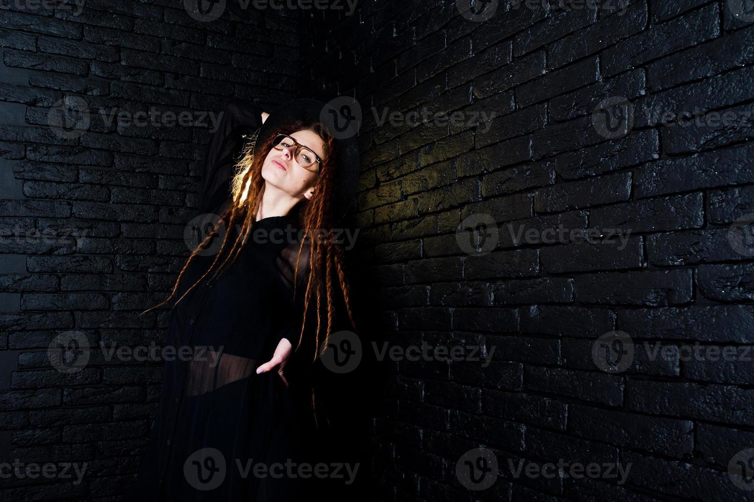 tournage en studio d'une fille en noir avec des dreads, des lunettes et un chapeau sur fond de briques. photo