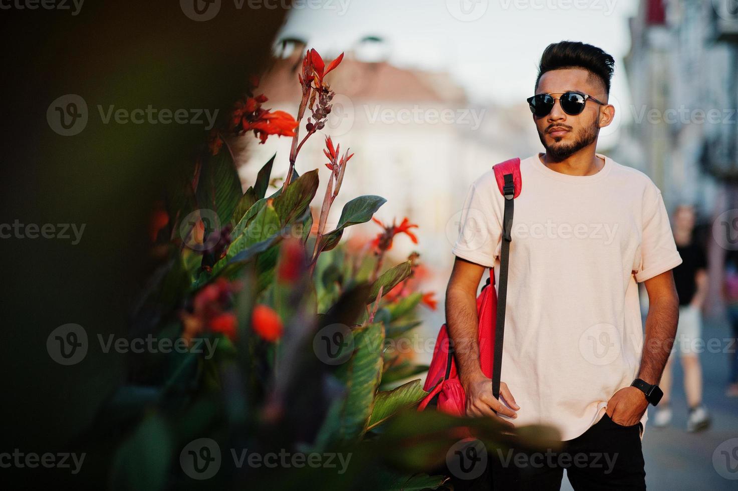 homme de barbe indien élégant au t-shirt rose, lunettes de soleil et sac à dos. modèle indien posé en plein air dans les rues de la ville du coucher du soleil. photo