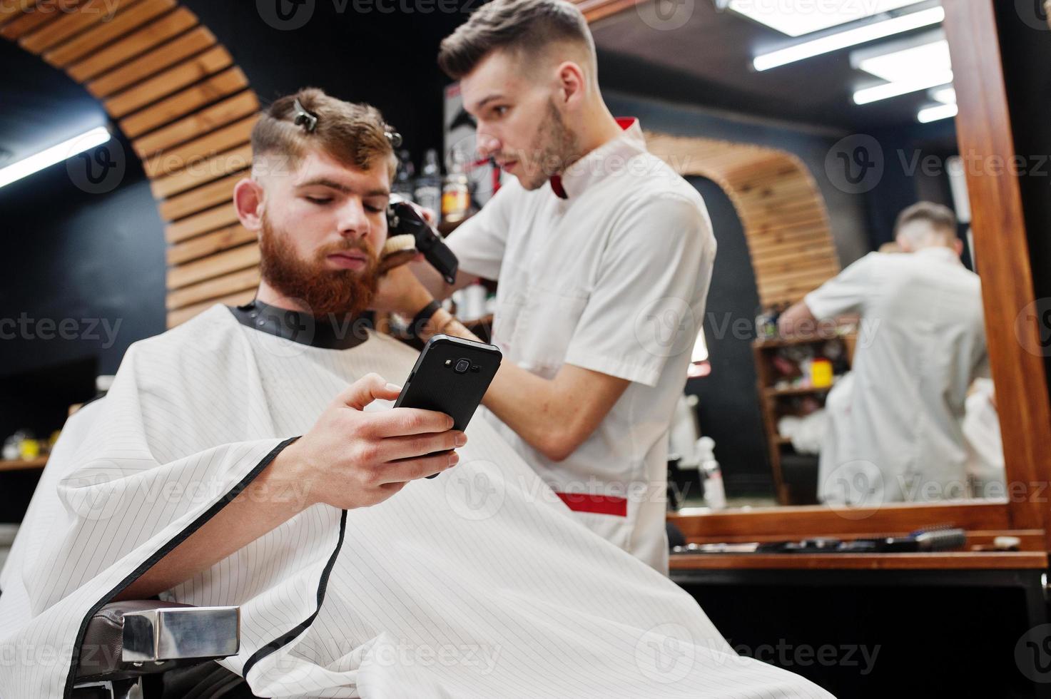 jeune homme barbu se coupe les cheveux par un coiffeur assis sur une chaise au salon de coiffure. âme de barbier. photo