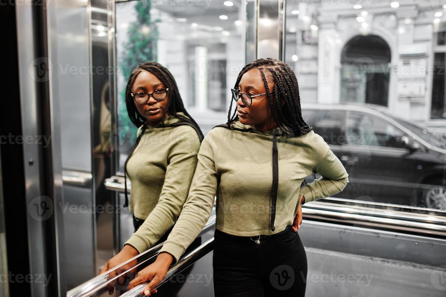 portrait d'une jeune femme positive à la peau foncée portant un sweat à capuche vert et des lunettes debout à l'ascenseur. photo