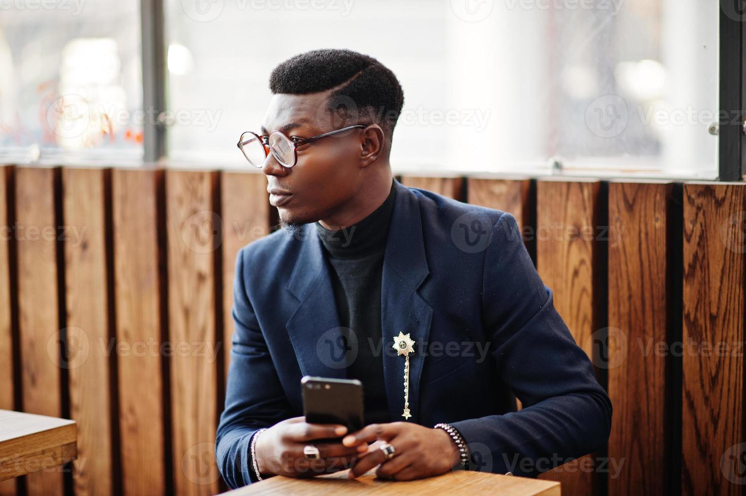 un homme afro-américain à la recherche étonnante porte un blazer bleu avec une broche, un col roulé noir et des lunettes posées dans la rue. gars noir à la mode assis derrière une table avec un téléphone portable. photo