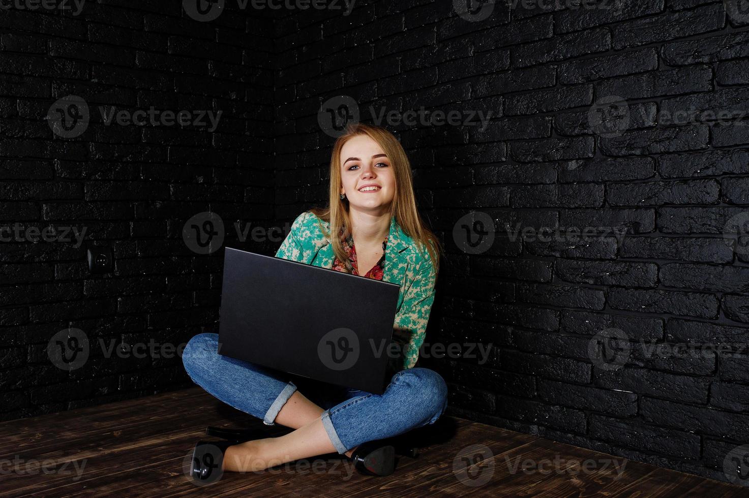 fille blonde élégante en veste et jeans avec ordinateur portable contre un mur noir en brique au studio. photo