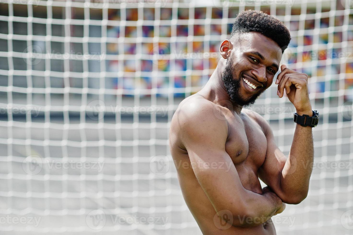 bel homme musclé torse nu afro-américain sexy à la salopette en jean posée à l'herbe verte contre les portes de football du terrain du stade. portrait d'homme noir à la mode. photo