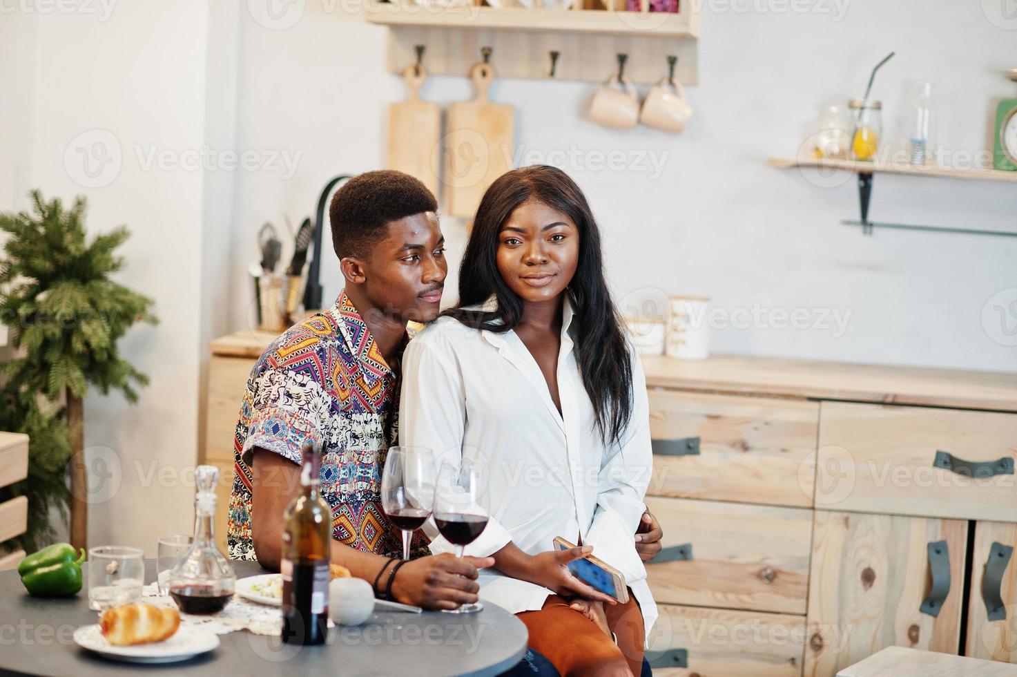 amoureux de couple afro-américain buvant du vin dans la cuisine à leur rendez-vous romantique avec un téléphone portable à portée de main. photo