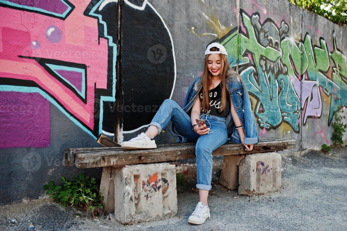 une fille hipster décontractée et élégante en casquette et en jean porte de la musique à partir d'un casque de téléphone portable contre un grand mur de graffitis avec une bombe. photo