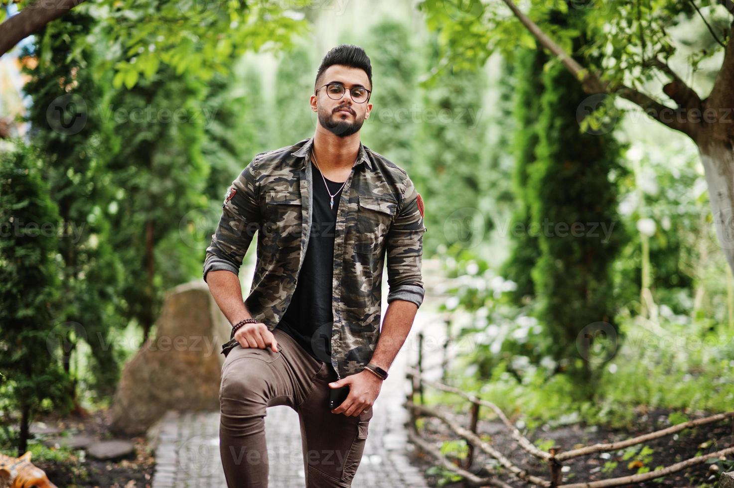 impressionnant beau grand homme macho à la barbe arabe dans des lunettes et une veste militaire posée à l'extérieur. photo