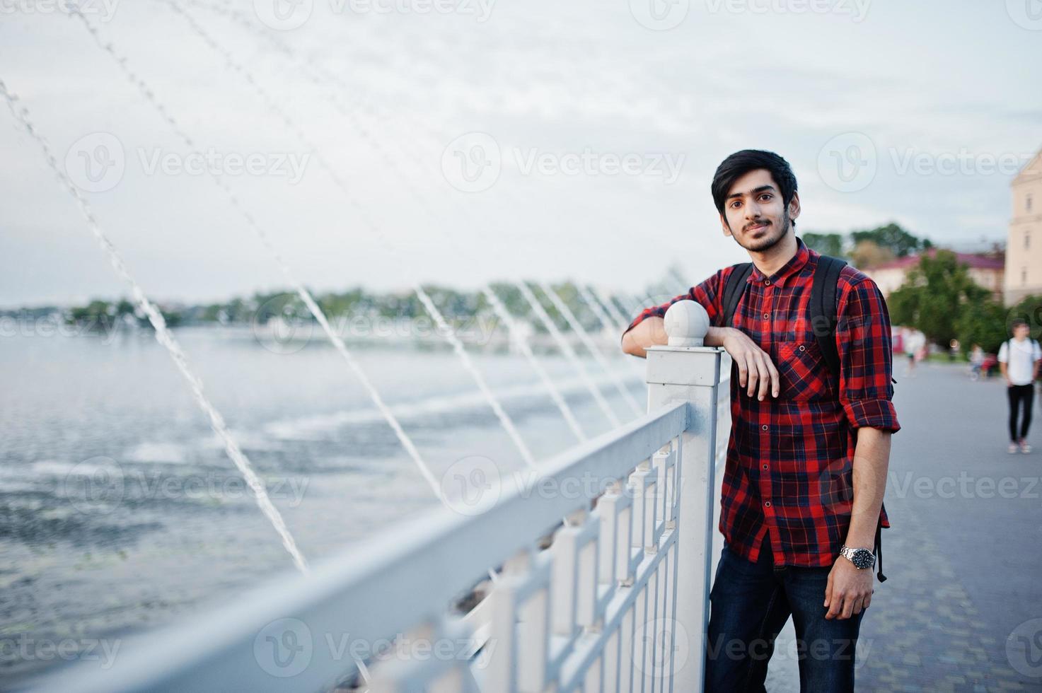 jeune étudiant indien en chemise à carreaux et jeans avec sac à dos posé sur la ville du soir contre les fontaines. photo