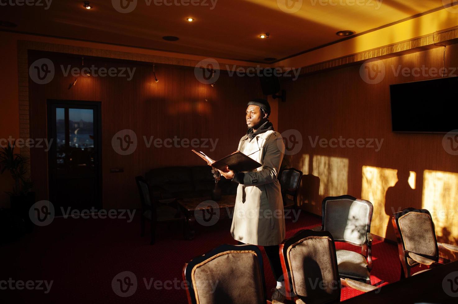 bel homme afro-américain posant à l'intérieur de la pièce avec des ombres de lumière du soleil en chapeau noir et manteau beige. photo