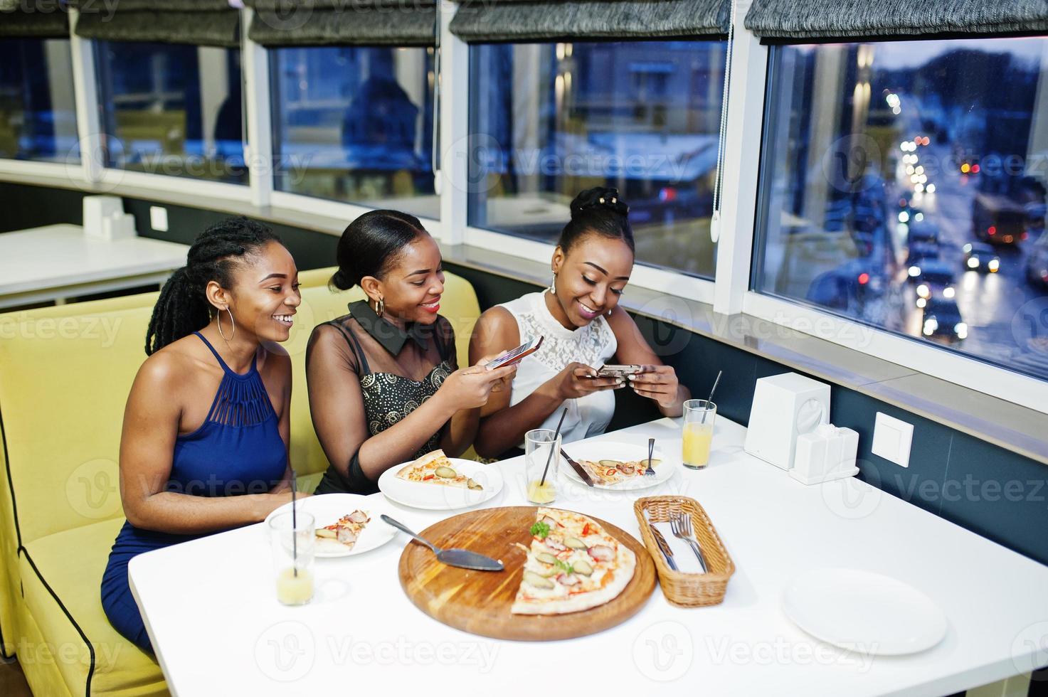 trois femmes africaines en robe posant au restaurant, mangeant de la pizza et buvant du jus, faisant des photos au téléphone.