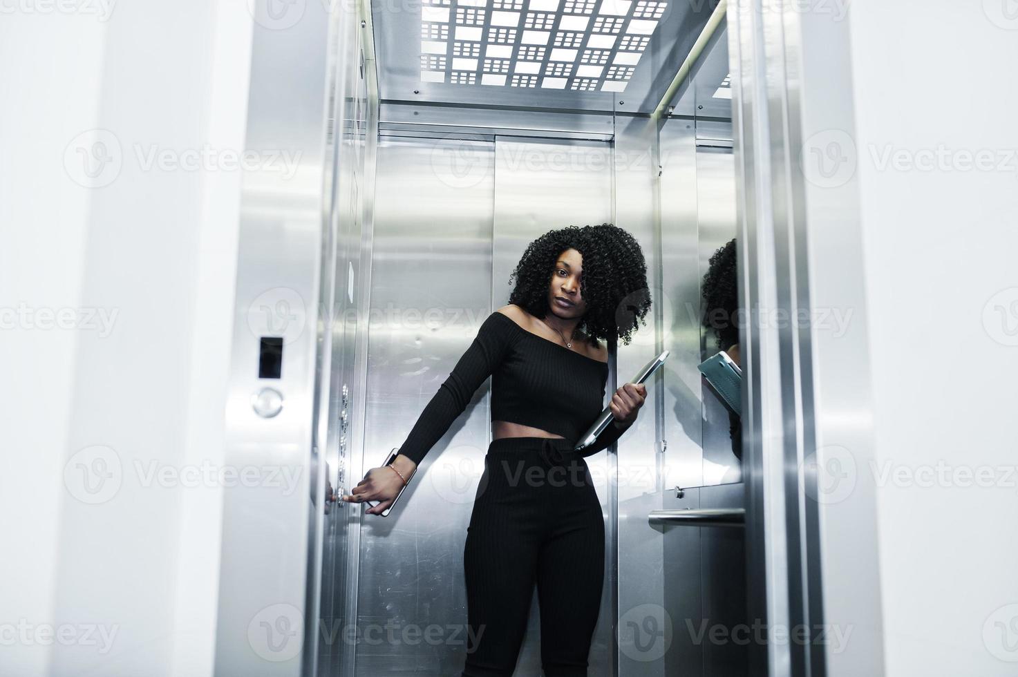 jeune belle femme d'affaires afro-américaine à la mode avec une coiffure afro en noir élégant, debout à l'ascenseur avec téléphone et ordinateur portable à portée de main. photo