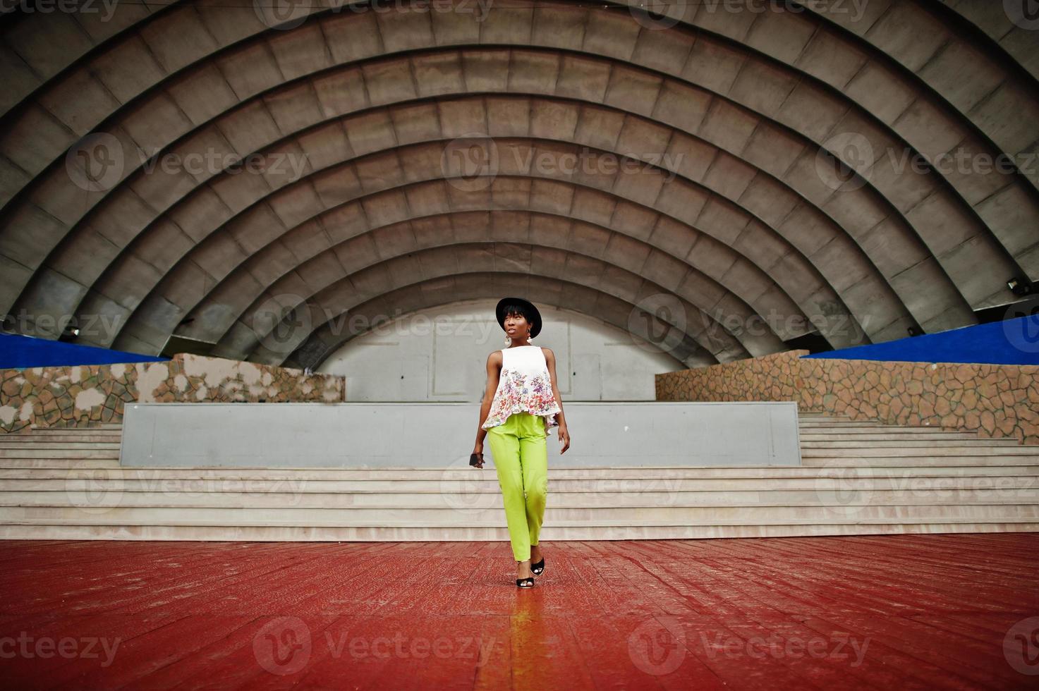 incroyable femme modèle afro-américaine en pantalon vert et chapeau noir posé en plein air contre le hall de l'arène. photo