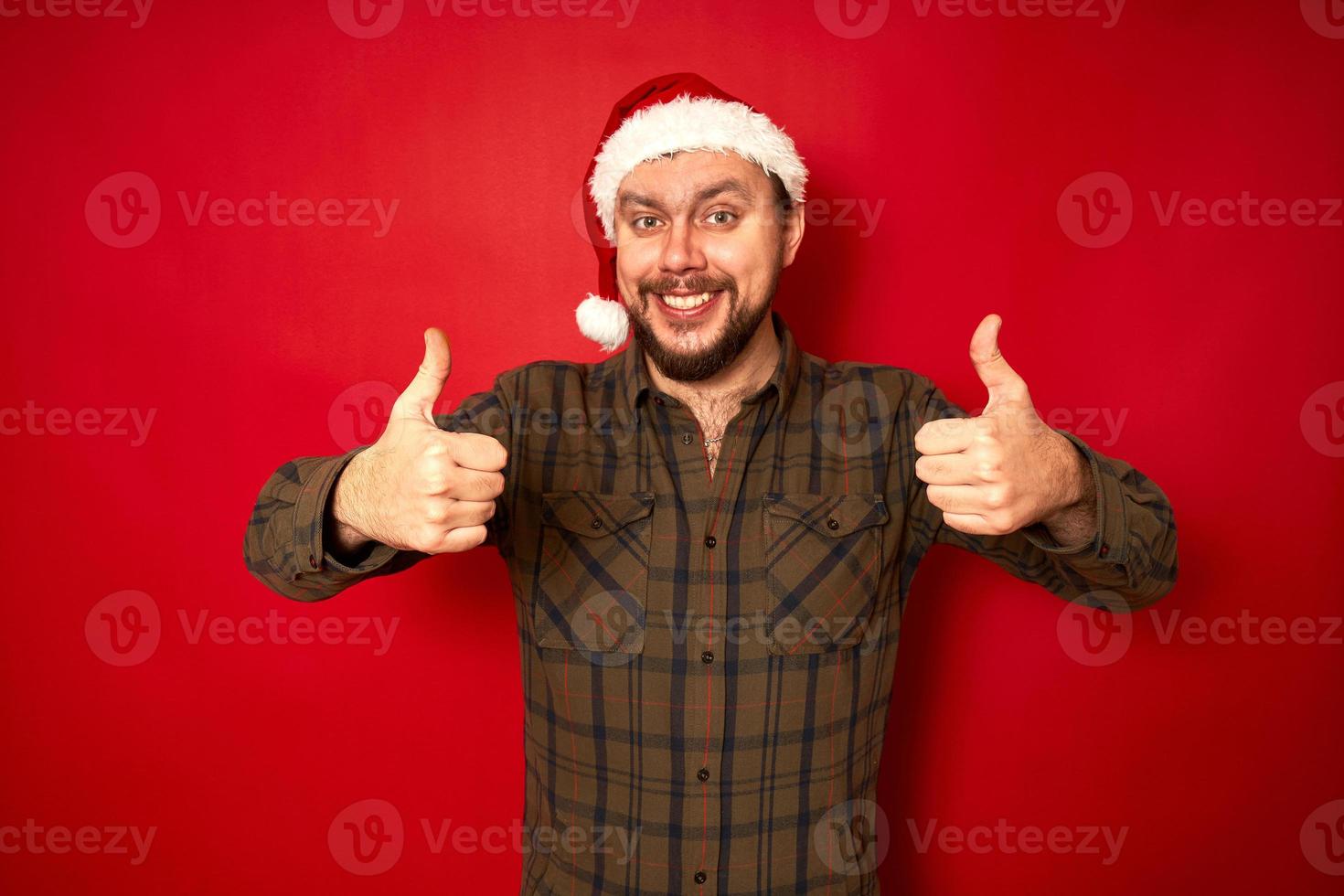 homme souriant au chapeau de noël, vêtements décontractés montre les pouces vers le haut avec les deux mains isolés sur fond de studio rouge avec un espace pour le texte. concept - vacances, nouvel an, promotion, remises, offre favorable photo