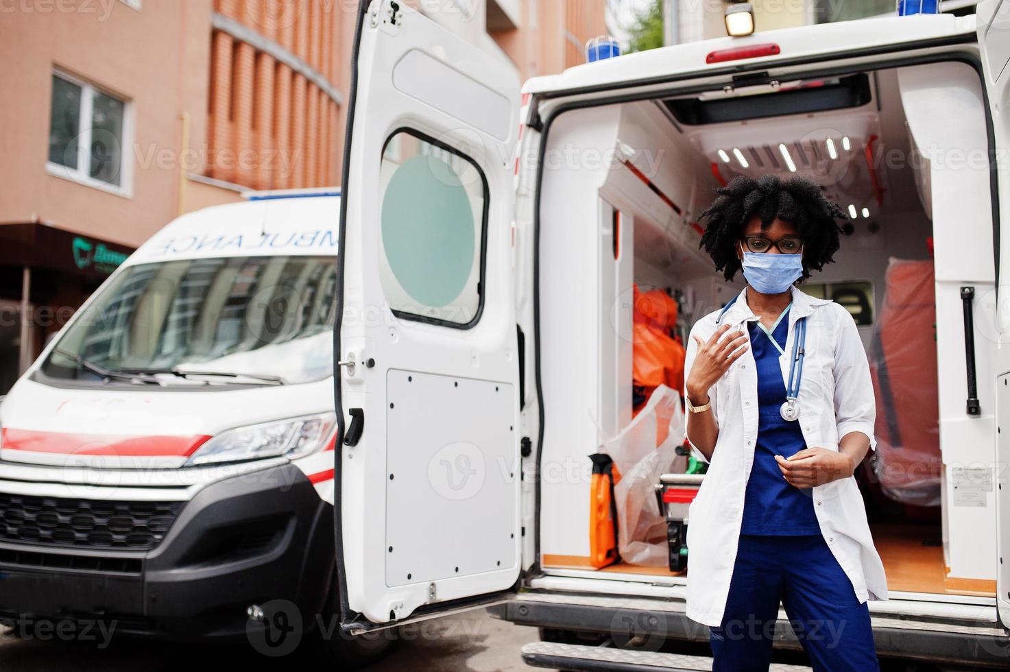 paramédic féminin afro-américain dans le masque médical de protection du visage debout devant la voiture d'ambulance. photo