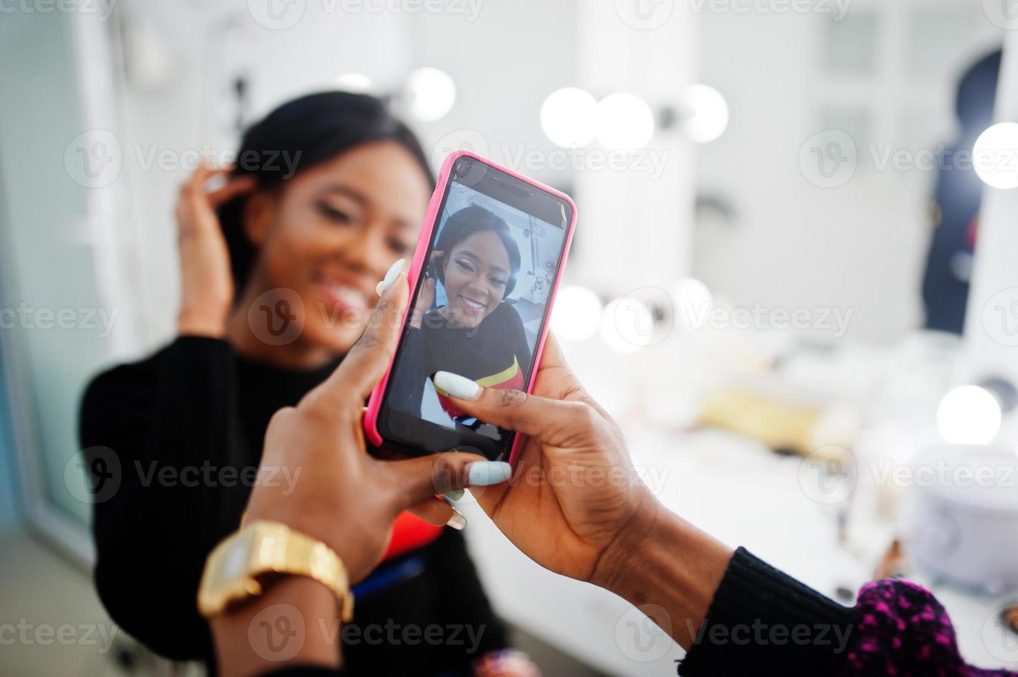 femme afro-américaine se maquillant par une maquilleuse au salon de beauté. l'artiste fait une photo sur son téléphone portable de son travail.