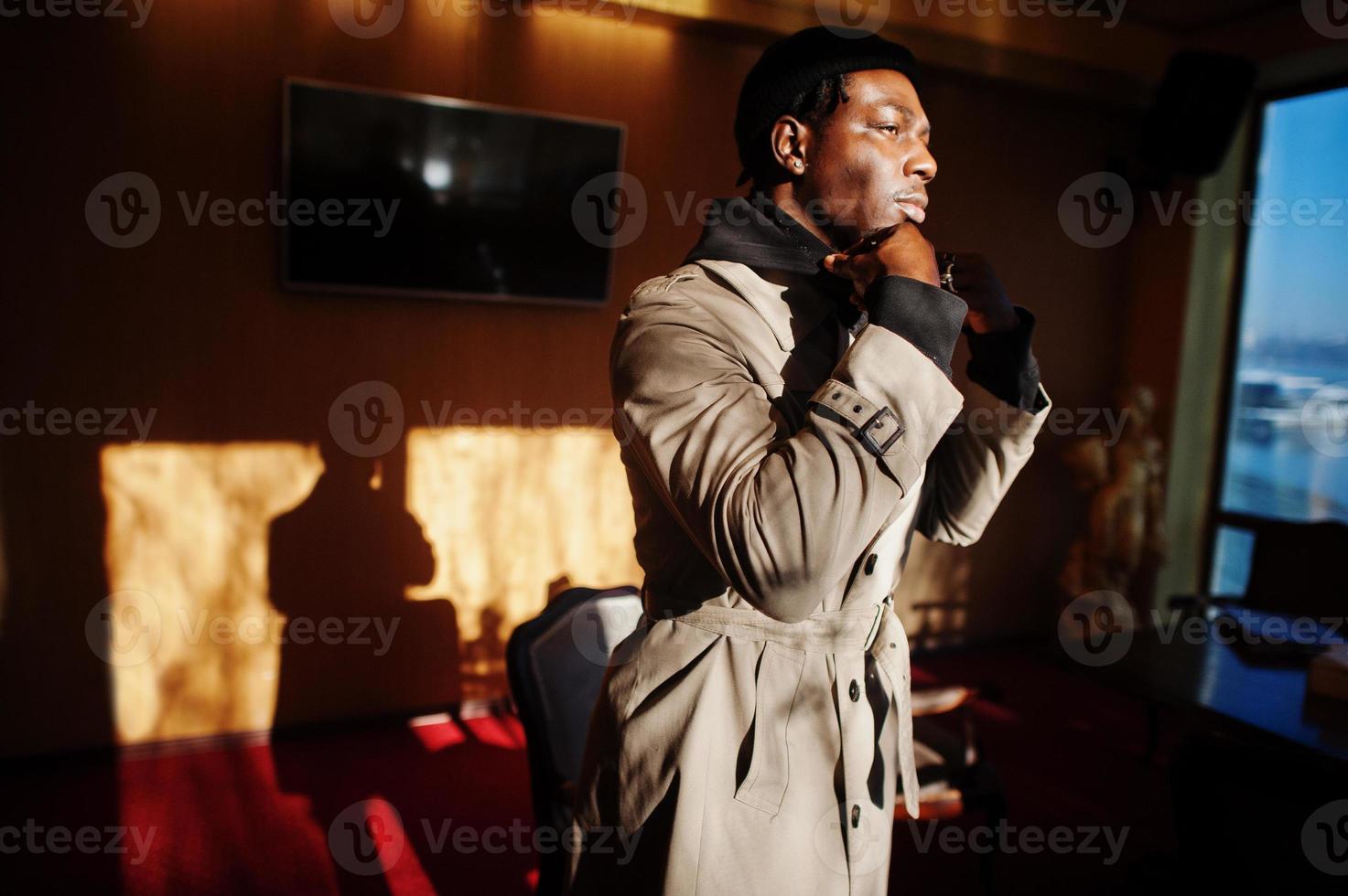 bel homme afro-américain posant à l'intérieur de la pièce avec des ombres de lumière du soleil en chapeau noir et manteau beige. photo