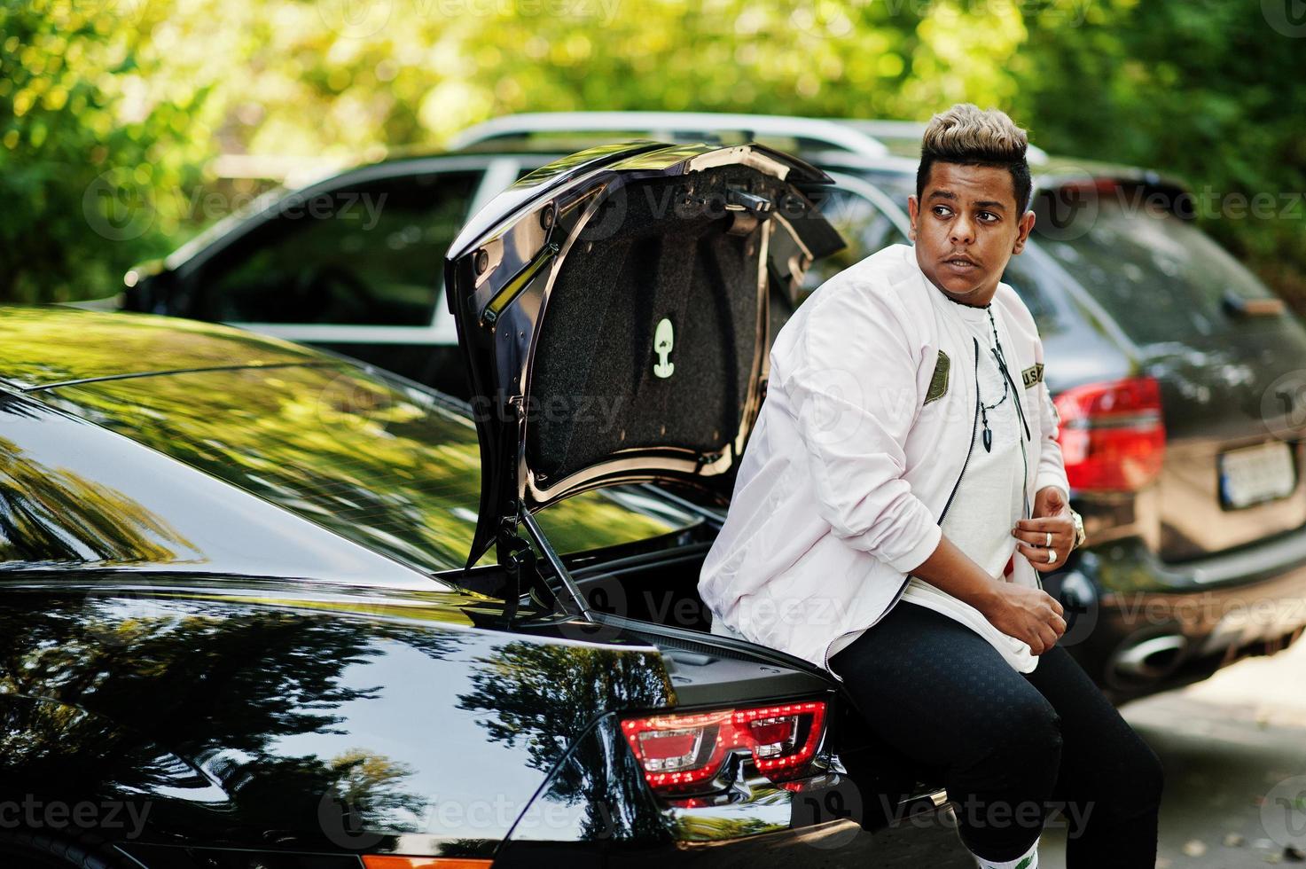 homme arabe hipster élégant posé en plein air dans la rue et assis sur le coffre de sa voiture de muscle noire. homme noir riche. photo