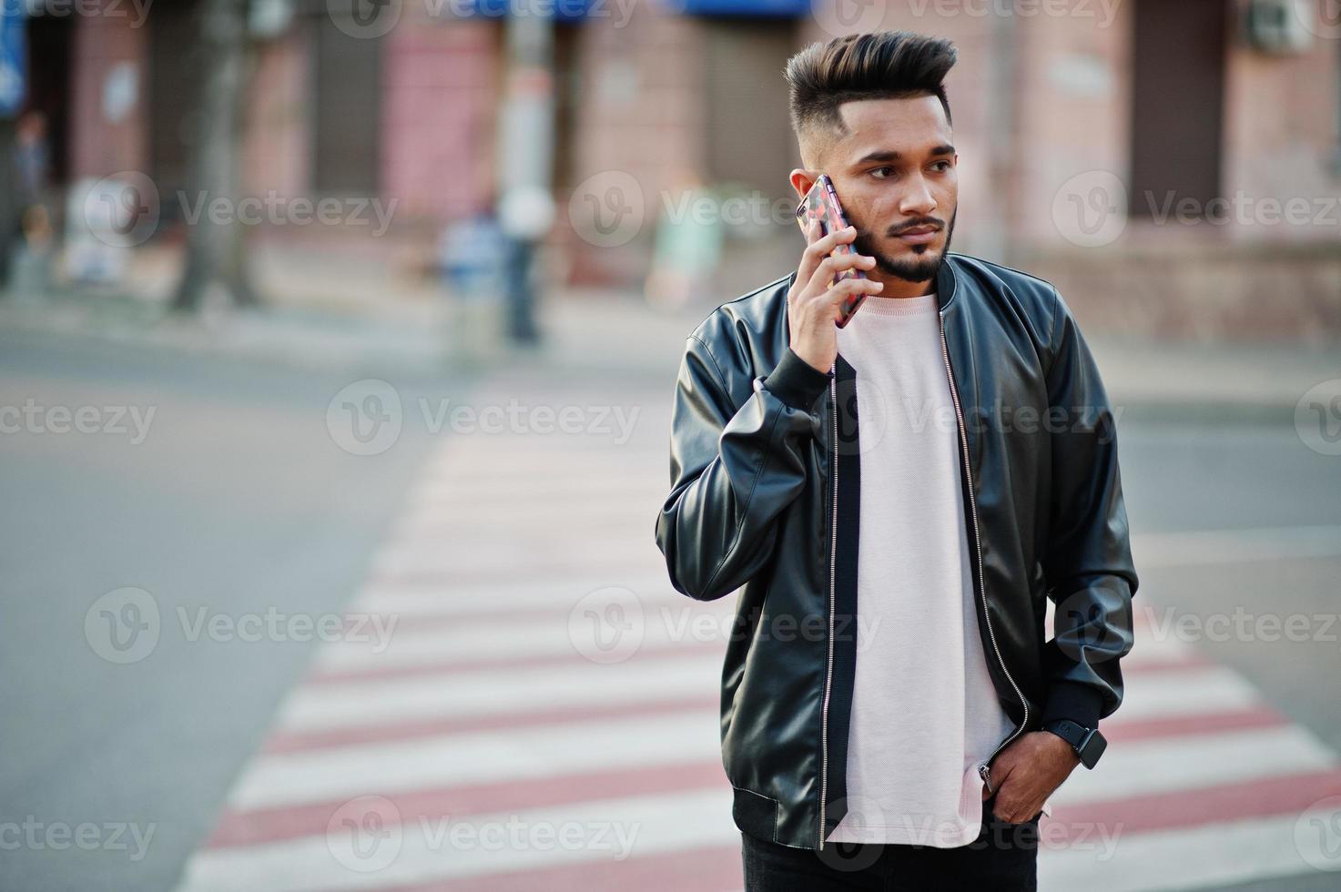 homme barbe indien élégant à la veste en cuir noir marchant sur le passage clouté. modèle indien posé en plein air dans les rues de la ville et téléphone parlant. photo