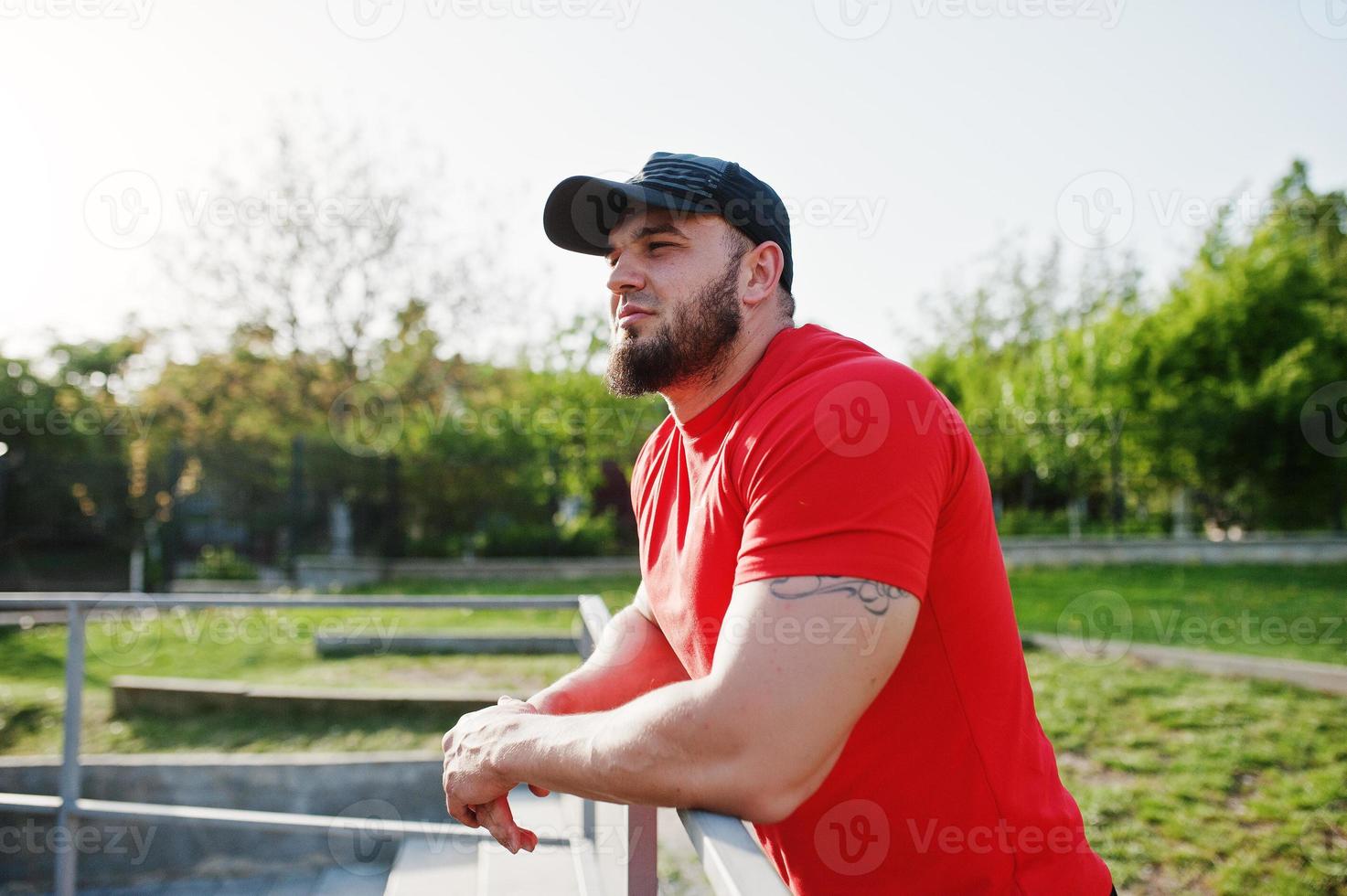 jeune homme musclé barbu brutal portant une chemise rouge, un short et une casquette au stade. photo