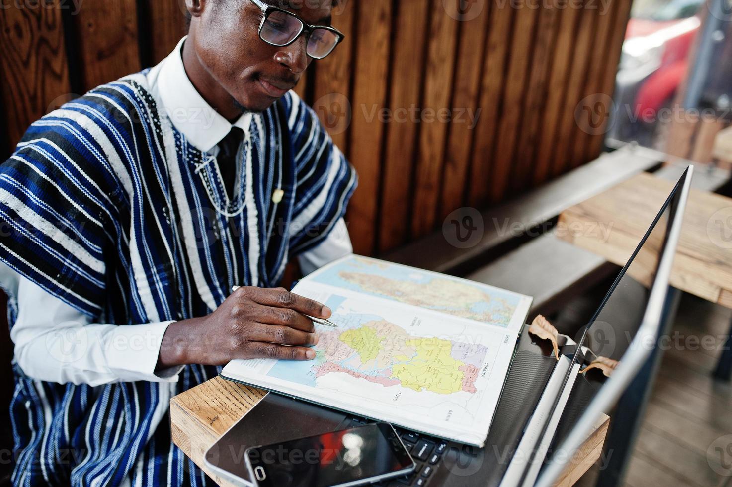 homme africain en vêtements traditionnels et lunettes assis derrière un ordinateur portable au café en plein air et regardant sur la carte de l'afrique et du ghana sur son ordinateur portable. photo