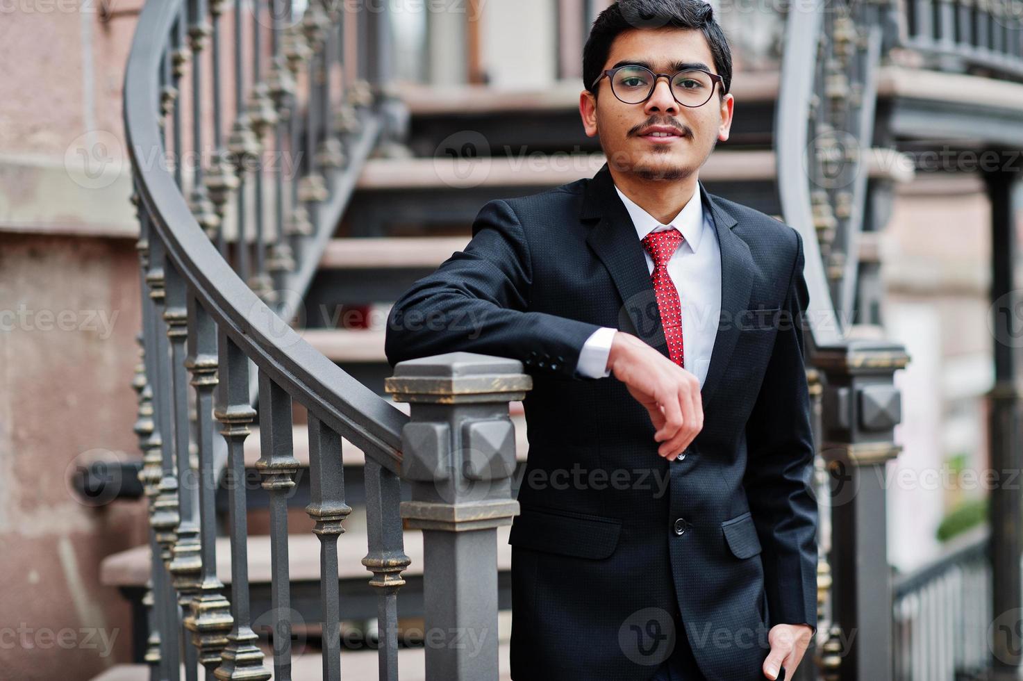 jeune homme indien à lunettes, porter un costume noir avec une cravate rouge posé à l'extérieur contre des escaliers en fer. photo