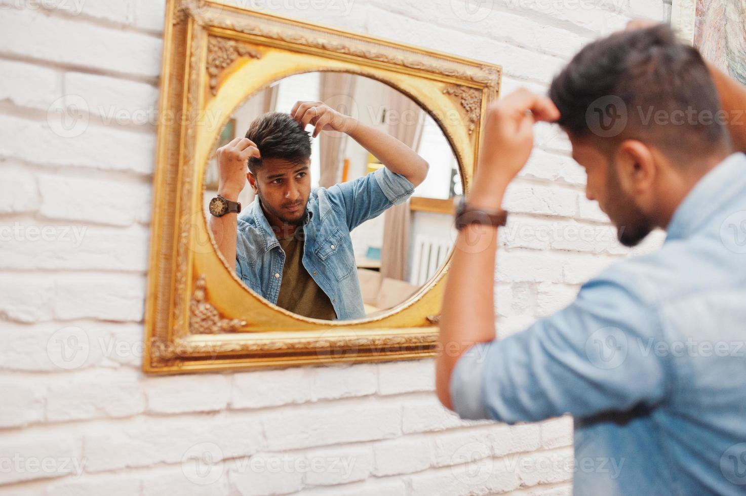 portrait d'un beau sud-asiatique barbu réussi, jeune pigiste indien en chemise de jeans bleu debout dans un café et regarde le miroir. photo