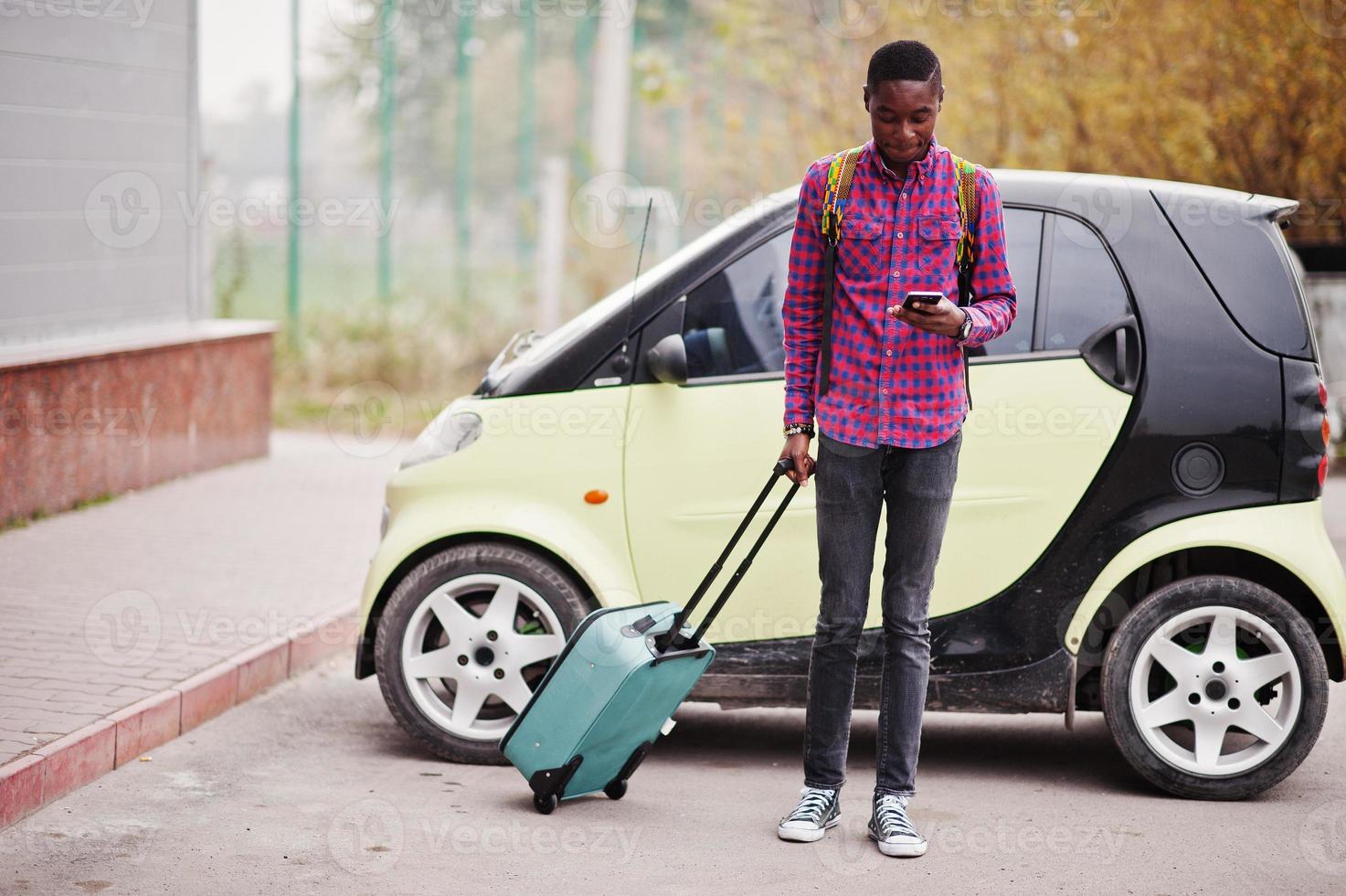 homme afro-américain en chemise à carreaux, avec valise et sac à dos. voyageur noir contre une petite voiture de ville. photo