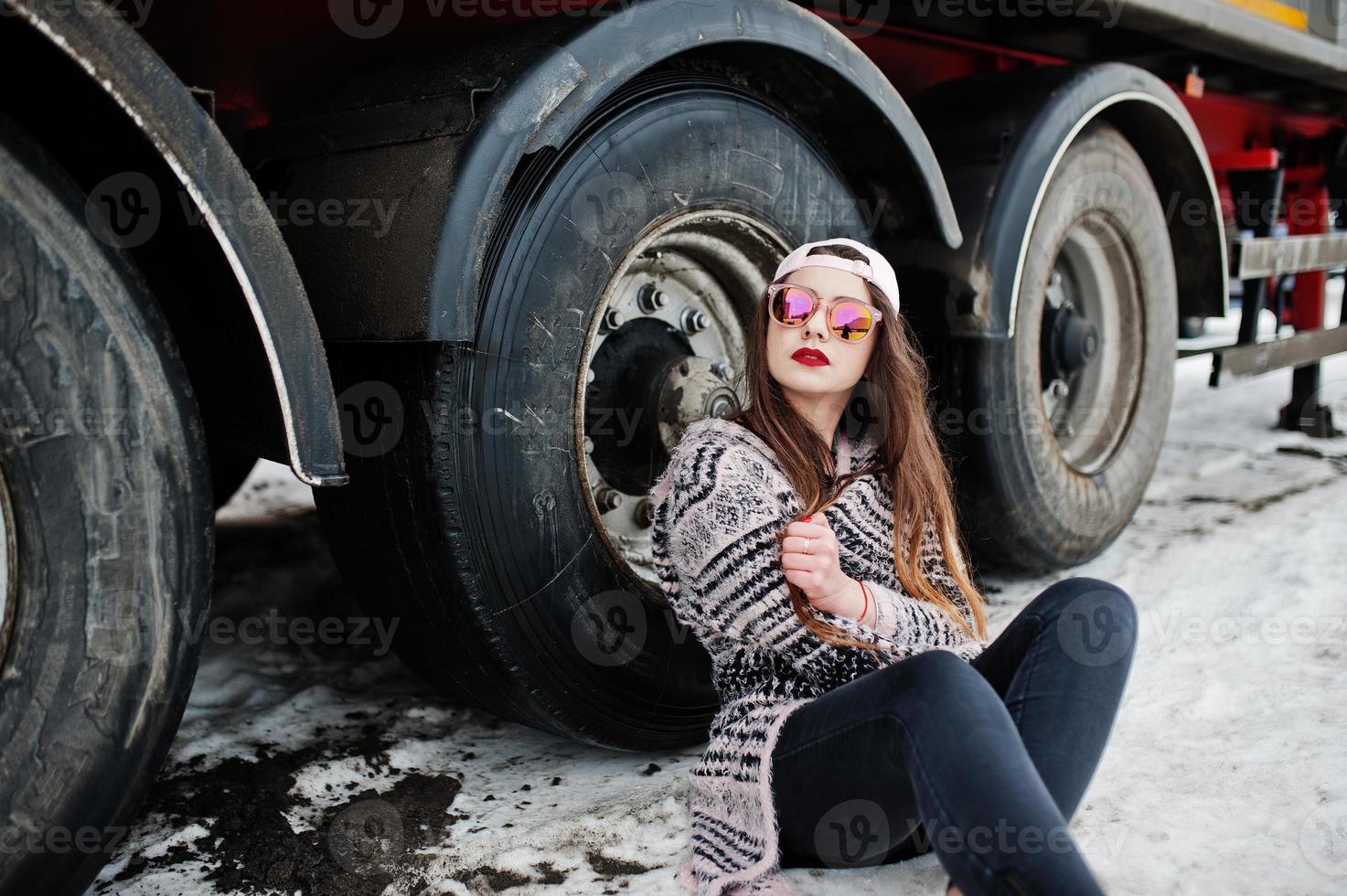 brune élégante fille décontractée en casquette et lunettes de soleil assise contre les roues du camion. photo