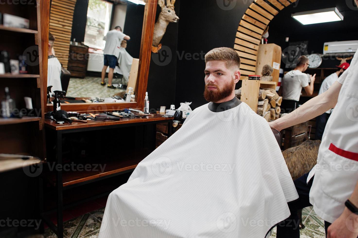 jeune homme barbu se coupe les cheveux par un coiffeur assis sur une chaise au salon de coiffure. âme de barbier. photo