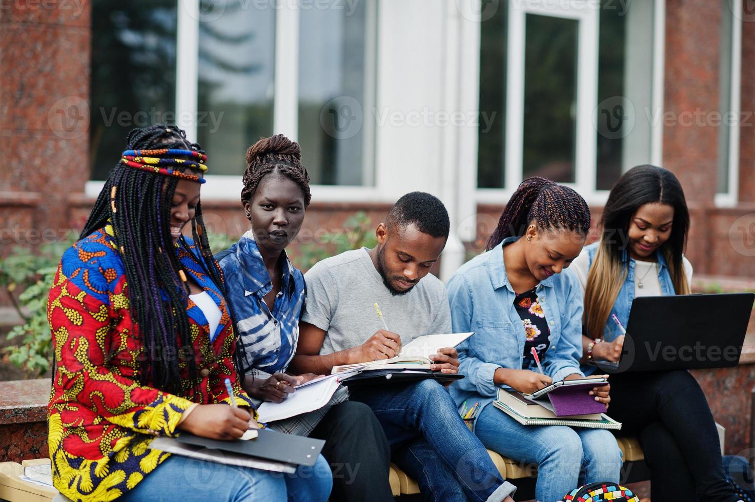 groupe de cinq étudiants africains qui passent du temps ensemble sur le campus de la cour universitaire. amis afro noirs étudiant au banc avec des articles scolaires, des cahiers d'ordinateurs portables. photo