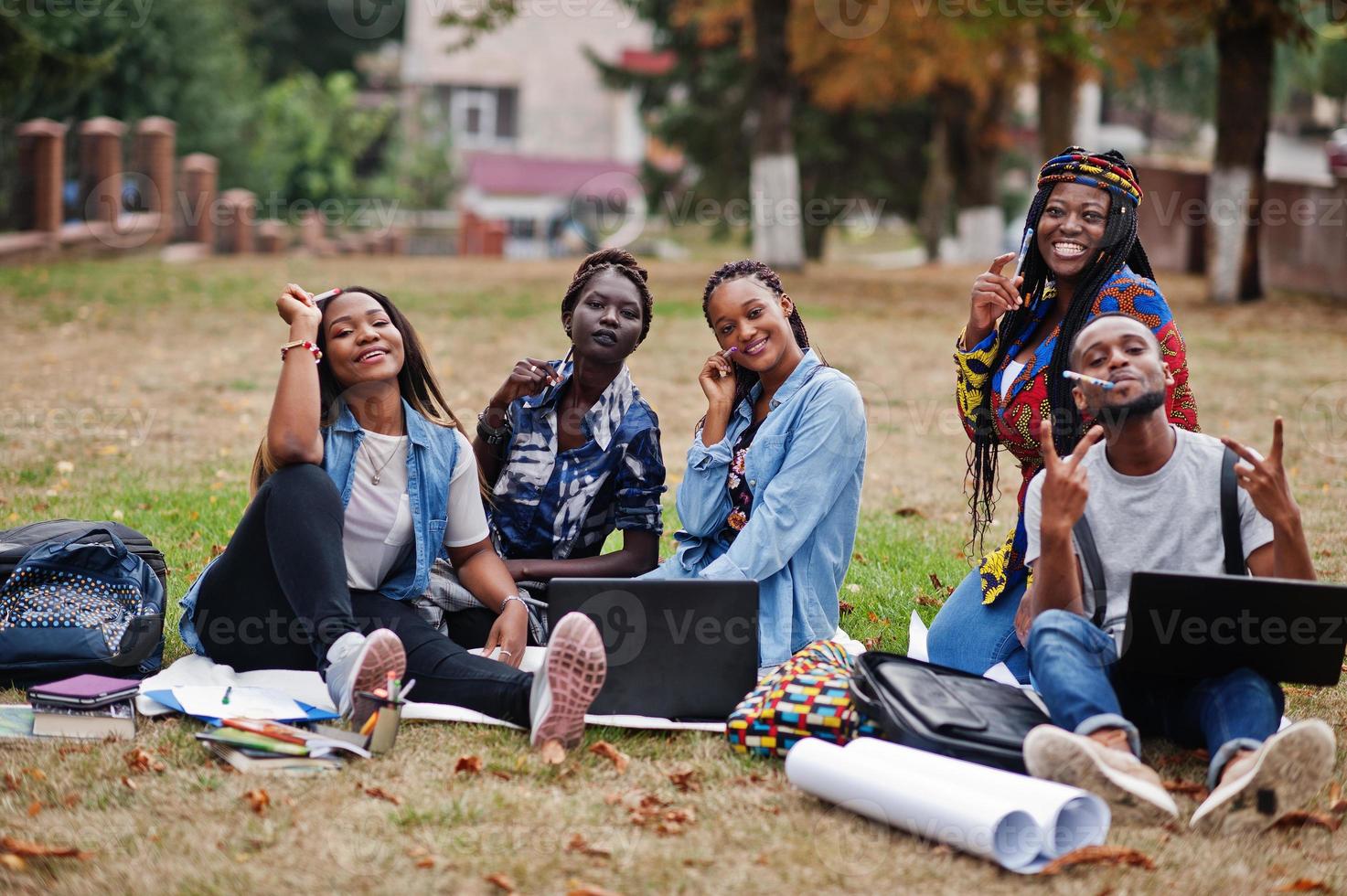 groupe de cinq étudiants africains qui passent du temps ensemble sur le campus de la cour universitaire. amis afro noirs assis sur l'herbe et étudiant avec des ordinateurs portables. photo