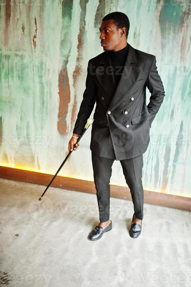 élégant gentleman afro-américain dans une élégante veste noire, tenant un bâton de marche rétro comme flacon de canne ou canne basculante avec poignée en boule de diamant doré. riche homme afro à la mode contre le mur vert. photo