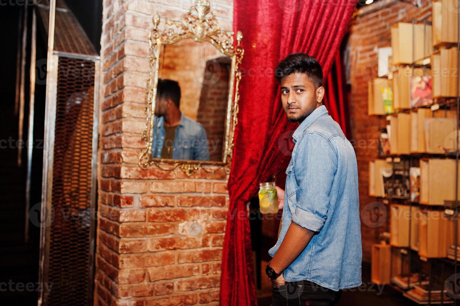 portrait d'un beau sud-asiatique barbu réussi, jeune pigiste indien en chemise de jeans bleu debout dans un café avec un cocktail contre le miroir. photo