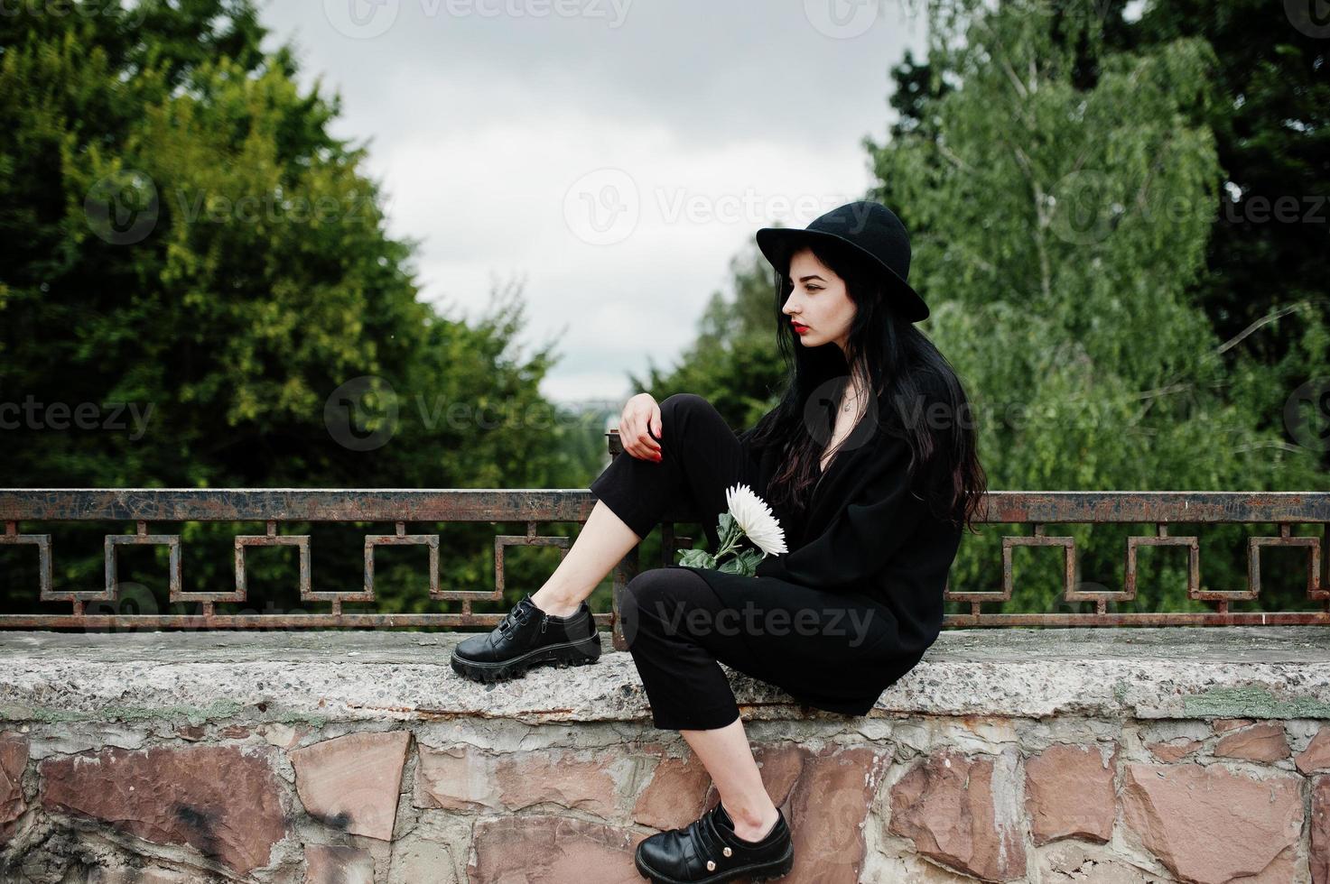 fille sensuelle tout en noir, lèvres rouges et chapeau. une femme dramatique gothique tient une fleur de chrysanthème blanche. photo