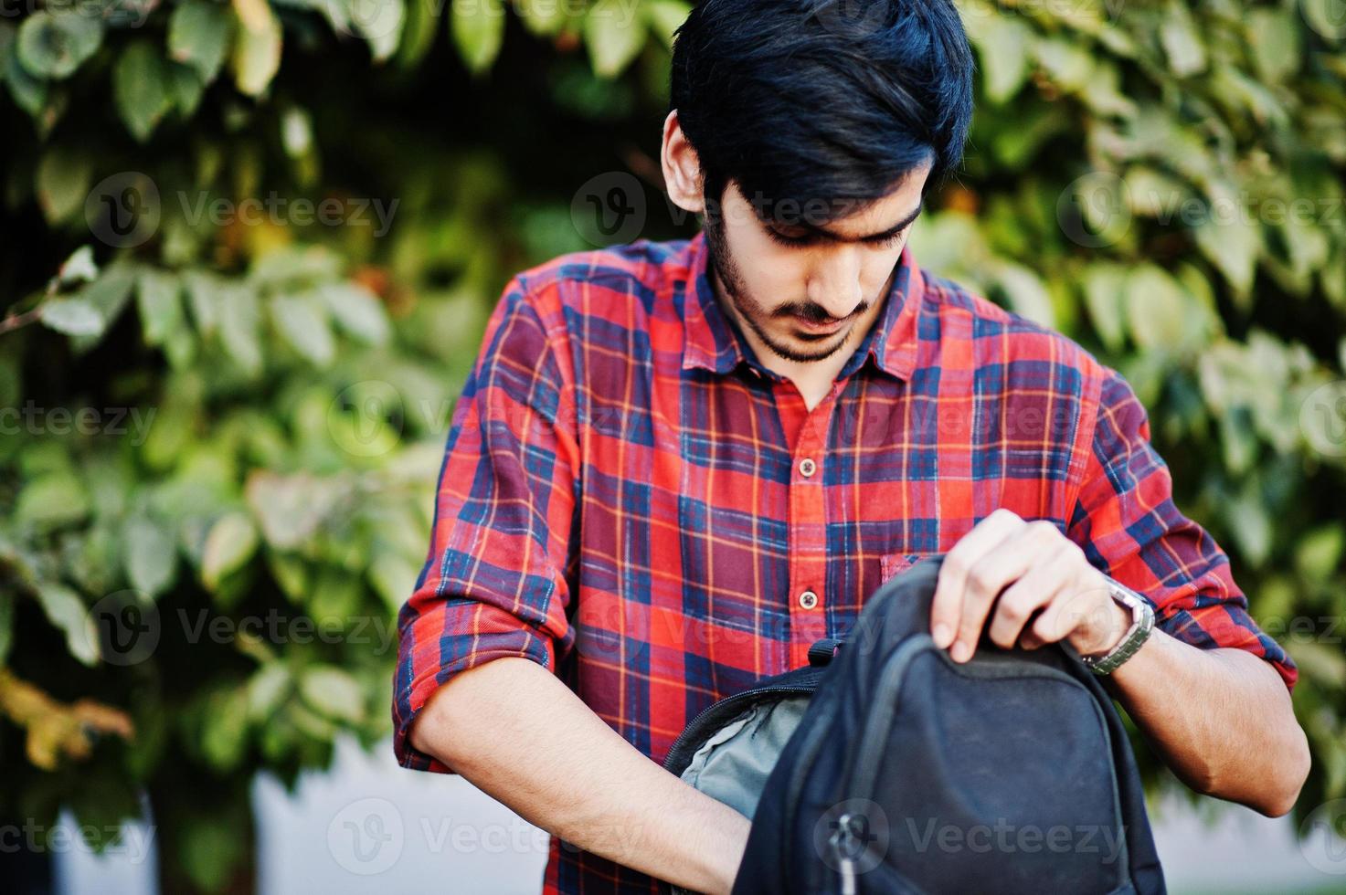 jeune étudiant indien en chemise à carreaux rouge et jeans avec sac à dos posé dans la rue. photo