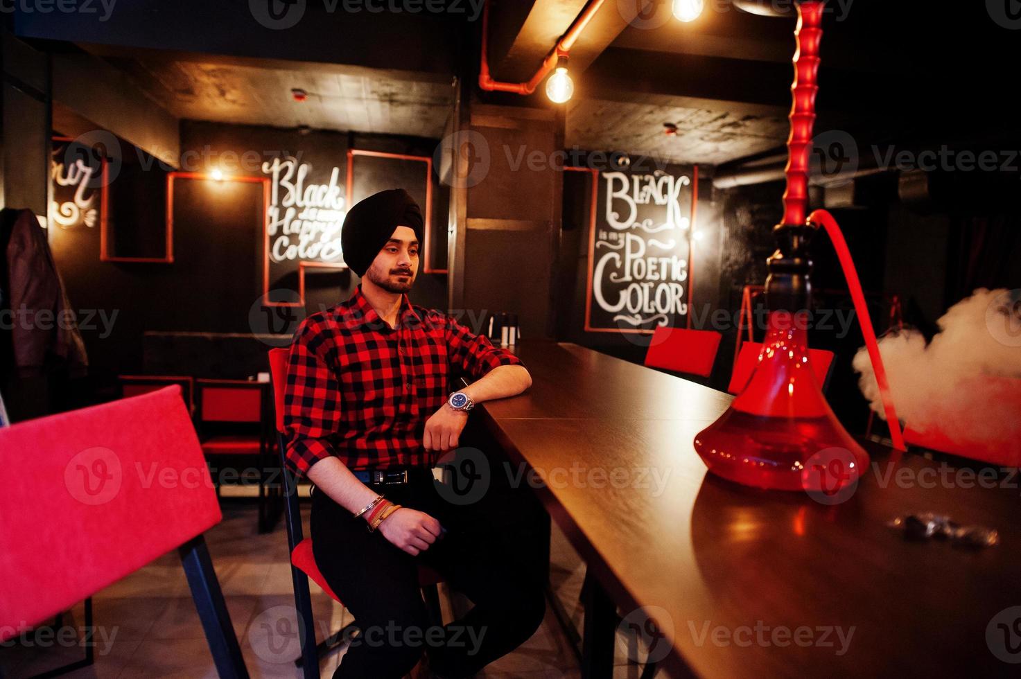 homme indien en chemise à carreaux et turban noir assis au bar avec narguilé. photo