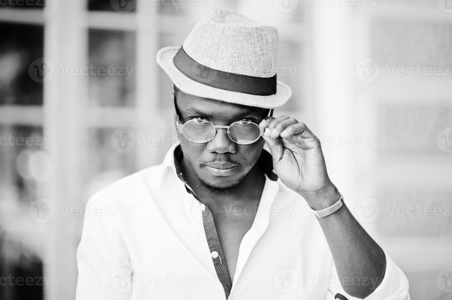 homme afro-américain élégant en chemise blanche et pantalon coloré avec chapeau et lunettes posés à l'extérieur. garçon modèle à la mode noir. photo