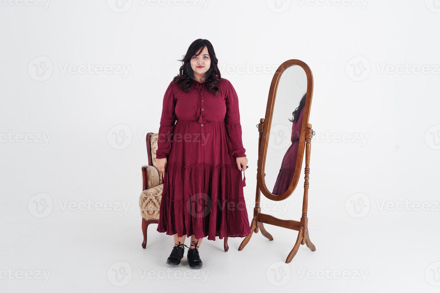 jolie femme sud-asiatique en robe rouge foncé posée au studio sur fond blanc contre le miroir et la chaise. photo