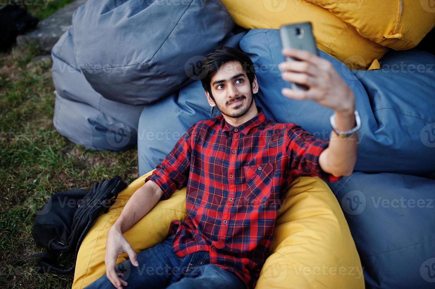 jeune étudiant indien en chemise à carreaux et jeans assis et se détendre sur des oreillers extérieurs et faire du selfie. photo