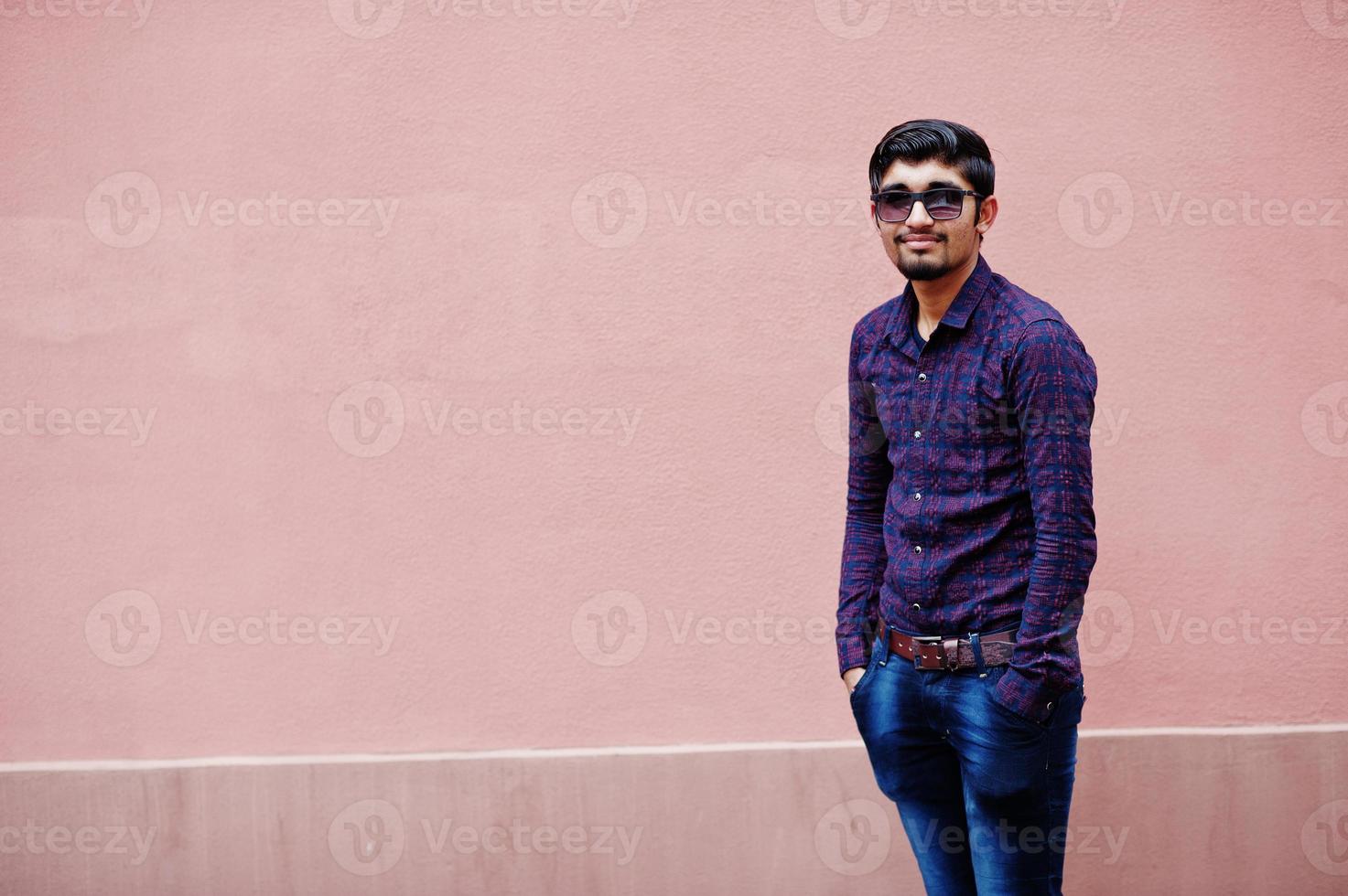 jeune homme indien en chemise et lunettes de soleil posé contre le mur rose. photo