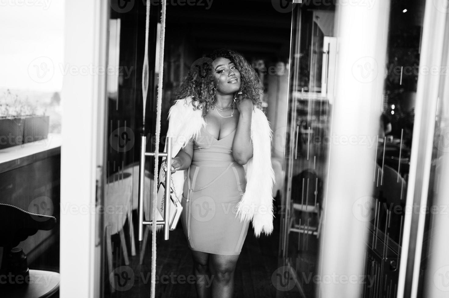 femme afro-américaine glamour en robe jaune et cape en laine blanche avec sac à main posé à l'entrée de la porte vitrée du restaurant. photo